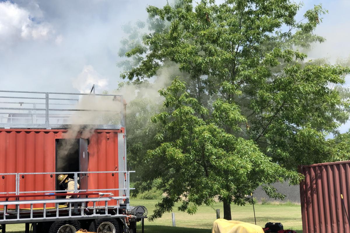 Rear door of red fire training trailer opened with smoke coming out next to a large green tree.