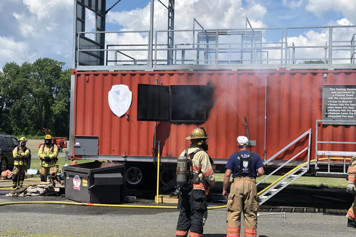 Two instructors conversing while watching the smoke escaping the red fire training trailer.
